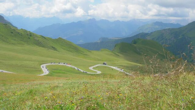 Scenic and winding mountain road through green meadows. In the background the Dolomites in a blue but cloudy sky. In the foreground, the rows of grass are moving by the wind. camera move to the left