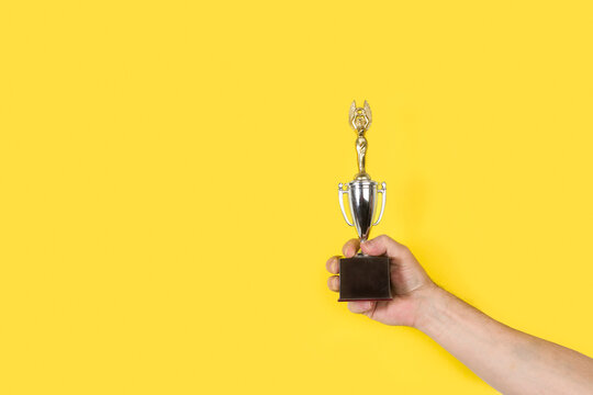 Mano de hombre sosteniendo y levantado un trofeo sobre un fondo amarillo liso y aislado. Vista de frente. Copy space. Concepto: Competici&oacute;n
