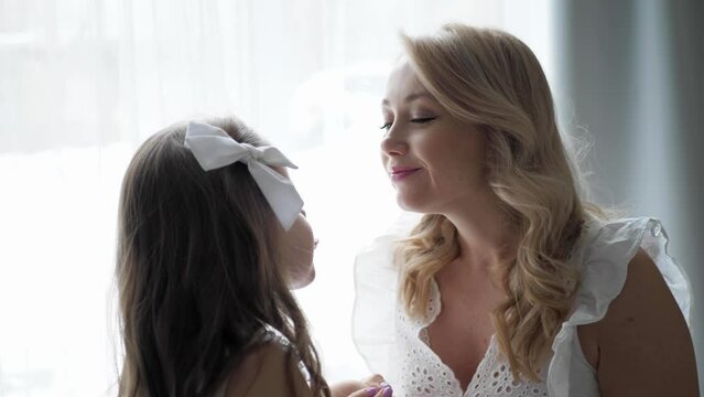 beautiful family mother and daughter in white dresses are sitting on the floor and kiss by a large window