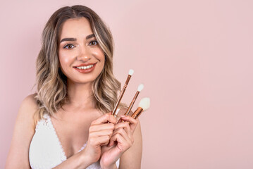 Beutiful blonde girl with natural makeup on light pink background, make up brushes in her hands, copy space