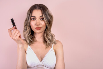 Fototapeta premium Beutiful blonde girl with natural makeup on light pink background, holding a lipstick , copy space