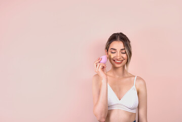 Fototapeta premium Beautiful blonde girl with natural makeup on light pink background, applying foundation on her face with a beauty blender, copy space