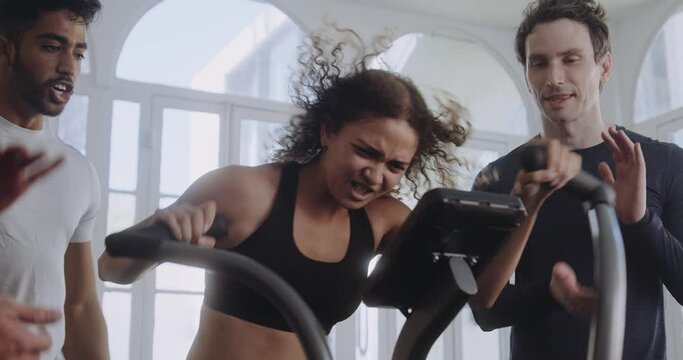 Biracial female working hard on the bike in a modern style gym. Friends motivating and cheering her on.