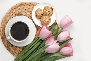 White cup of coffee with pink tulips on white background. Morning coffee on wicker tray. View from above of morning coffee, cookies and flowers. Flat lay. Breakfast in bed. Stylish home interior decor