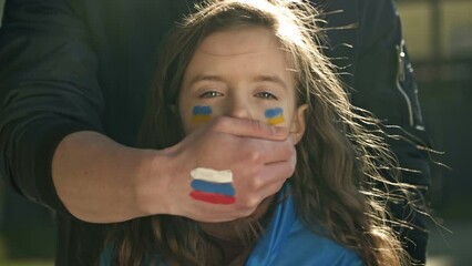 Man's hand with the image of the Russian flag roughly closes the mouth of a little girl with the Ukrainian flag. Children's tears of war. Freedom for Ukraine. Russian aggression.