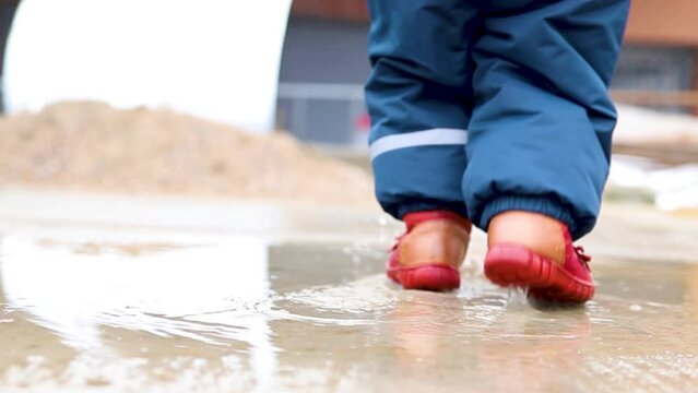 The kid walks through a puddle in the spring season. Children's feet in red boots squelching in a puddle.