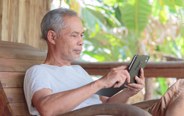Asian senior man on a rocking chair using a smartphone tablet to surf the Internet and read online news. Technology and a relaxed retirement lifestyle