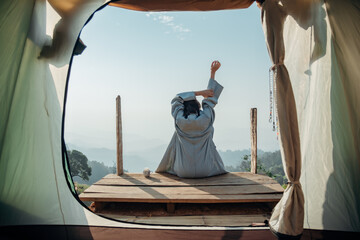 Rearview image of a woman sitting on a wooden balcony while watching a beautiful mountain and nature view outside the tent with her cup of coffee.