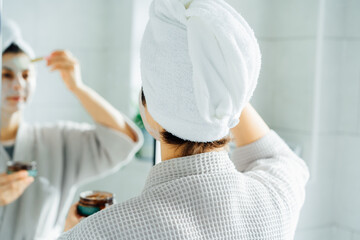 Woman in bathrobe with a towel on her head looking in the mirror and applying facial cosmetic clay...