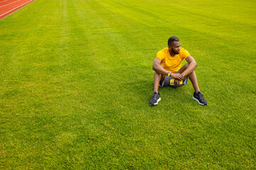 Fototapeta na wymiar Concentrated athletic man in stylish sportswear sitting at the stadium stretching legs. Sportive african american male working out, doing warm up before training outdoors concept