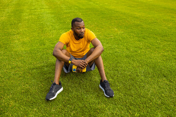 Fototapeta na wymiar Young adult male sitting on the grass in stylish sportswear after workout, hold a smartphone, relaxing. Sportive african american male taking a break from exercising, smiling, training outdoor concept