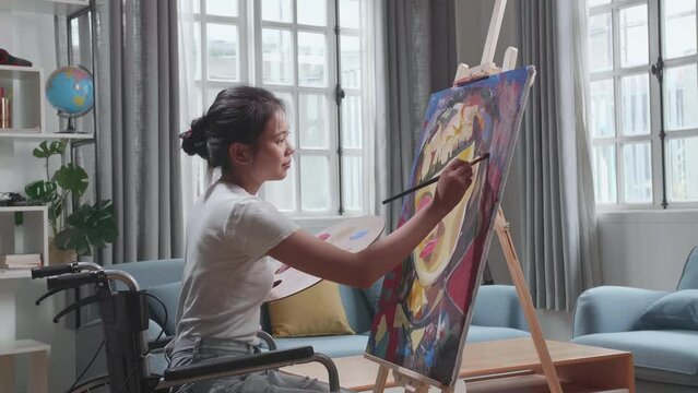 Side View Of An Asian Artist Girl In Wheelchair Holding Paintbrush Mixed Colour And Painting A Girl On The Canvas
