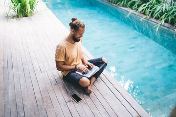 Millennial male with laptop computer working on online project browsing websites near pool at villa, software programmer communicating in social networks while creating web design via netbook app