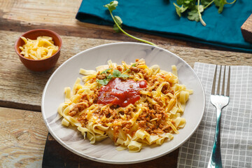 Pasta bolognese garnished with greens and cheese in a plate on a wooden table next to a fork and greens on a blue napkin.