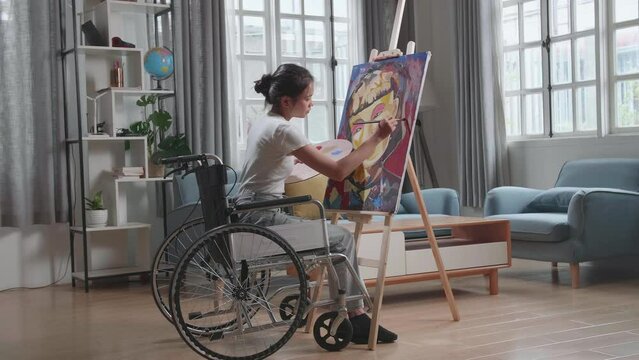 Side View Of An Asian Artist Girl In Wheelchair Holding Paintbrush Mixed Colour Thinking And Painting A Girl On The Canvas
