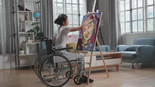 Talented Female Artist Sitting On Wheelchair, Enthusiastically Using Paint Brush She Creates With Oil Painting. Creative Studio With Canvas 
