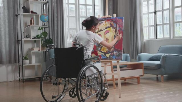 Back View Of An Asian Artist Girl In Wheelchair Holding Paintbrush Mixed Colour And Painting A Girl On The Canvas
