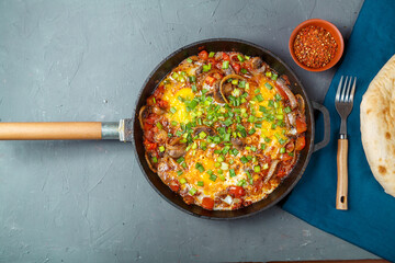 Shakshuka in a frying pan on a gray background next to a fork and spices on a blue napkin.