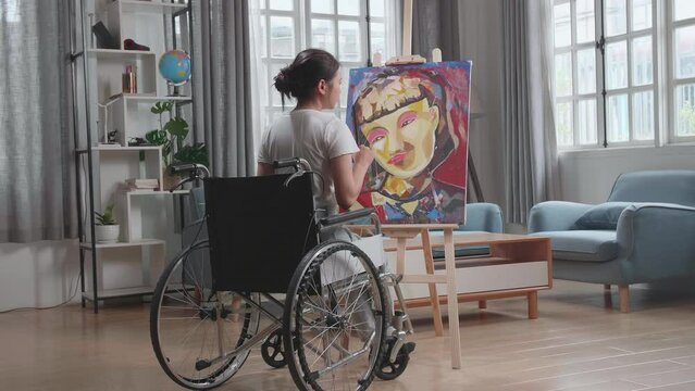 Back View Of An Asian Artist Girl In Wheelchair Holding Paintbrush Mixed Colour Thinking And Painting A Girl On The Canvas
