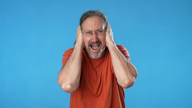 Displeased irritated sad angry elderly gray-haired bearded man 50s 60s wears orange shirt cover ears do not want to listen scream isolated on solid blue background studio portrait