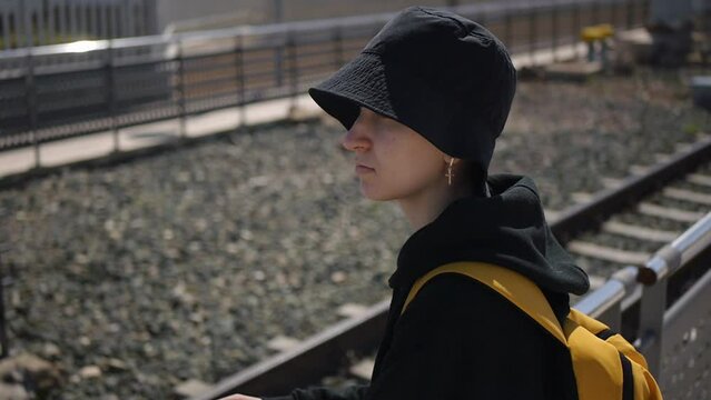 a teenage girl in black hat with yellow backpack standing near the railway. Refugee concept. Medium shot