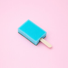 Kitchen sponge on a wooden popsicle stick isolated on a pastel blue background. Creative summer concept. Minimal hygiene or cleaning work idea.