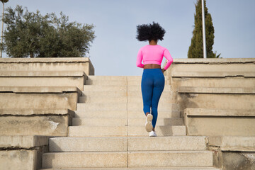 African-American woman with afro hair and sportswear, with fluorescent pink t-shirt and leggings, running up and down some stairs outdoors. Fitness concept, sport, street, urban.