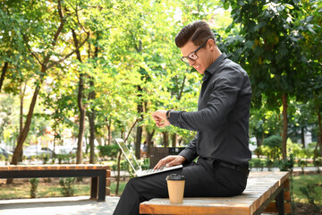 Handsome businessman with laptop looking at wristwatch in park