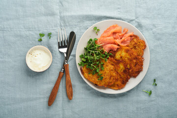 Potato pancakes. Fried homemade potato pancakes or latkes with cream, green onions, microgreens, red salmon and sauce in rustic plate on blue linen tablecloth. Rustic style. Healthy food. Top view.