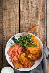 Potato pancakes. Fried homemade potato pancakes or latkes with cream, green onions, microgreens, red salmon and sauce in rustic plate on old wooden table background. Rustic style. Top view.