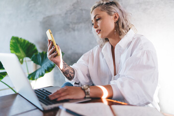 Young female freelancer with netbook reading received web message using mobile phone indoors,...