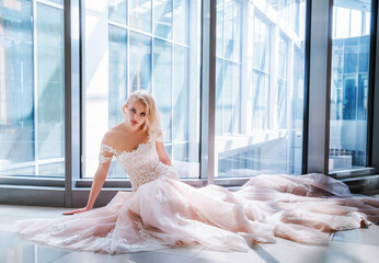 beautiful blonde bride in a luxurious wedding dress is sitting close to the big window