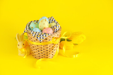 Wicker basket of painted Easter eggs, flowers and toy bunny on yellow background