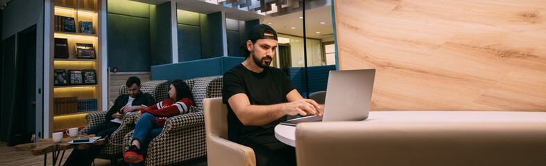 Men and women work in coworking. Young guy with a laptop at work online at the table and a couple of colleagues chatting, sitting on armchairs in a stylish room