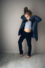 funny portrait of a fashionable girl child in a large gray jacket stands against gray wall next to...