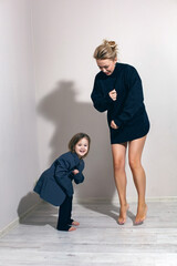 funny portrait of a fashionable girl child in a large gray jacket stands against gray wall next to...