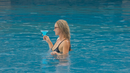 Middle shot pretty woman drinking blue cocktail alcohol liquor in swimming pool at hotel. Portrait of sexy girl in pool outdoor. Beautiful caucasian women with blond hair, black swimsuit, sunglasses.