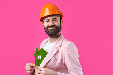 Portrait of young construction engineer wear orange hard hat, in a pink jacket standing on red studio background. A man holds a green eco house.