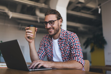 Cheerful programmer in classic eyeglasses watching received video on laptop computer and smiling, happy man in spectacles holding coffee cup and reading funny content publication browsed on netbook
