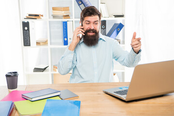 Happy guy talking on mobile phone working on laptop in office, business call
