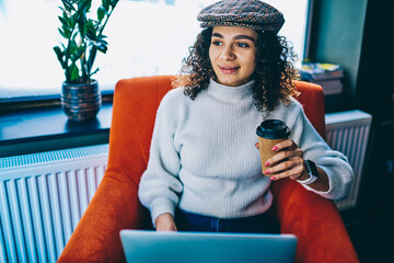 Millennial female in stylish hat holding takeaway caffeine beverage spending daytime for networking...