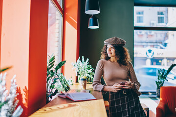 Millennial female teenager in trendy hat using 4g connection on modern cellular gadget - e learning in coworking with laptop device and education textbook, concept of technology and communication