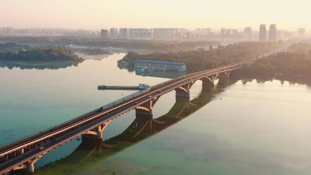 Amazing cinematic aerial view of cars and train on bridge over amazing Dnepr river and idyllic sunrise Kyiv panorama.