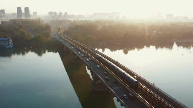 Cars and trains cross Dnepr river over stunning bridge, idyllic aerial flyover shot of summer sunrise Kyiv, Ukraine.
