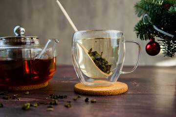 A transparent mug with tea and a wooden spoon stands on a wooden stand, there is a transparent teapot next to it and tea leaves are scattered on a dark background. High quality photo