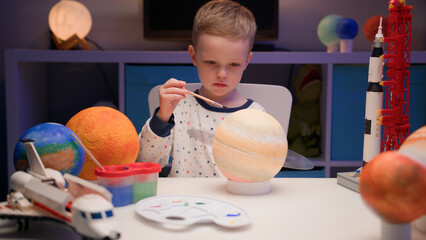 Blond boy paint planet solar system Saturn with colorful paint sitting home table in evening,...