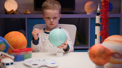 Blond boy paint planet solar system Uranus with colorful paint sitting home table in evening,...