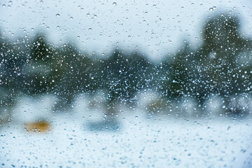 Raindrops on a window with the background of blurry green trees and snow