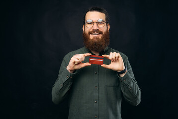 Ecstatic bearded man is holding his red credit card with both hands.