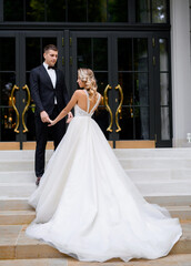 Side view of happy groom, wearing in glamour black suit, holding by hands with bride, smiling and looking to her while standing near modern building outdoor. Back view of elegant faceless woman